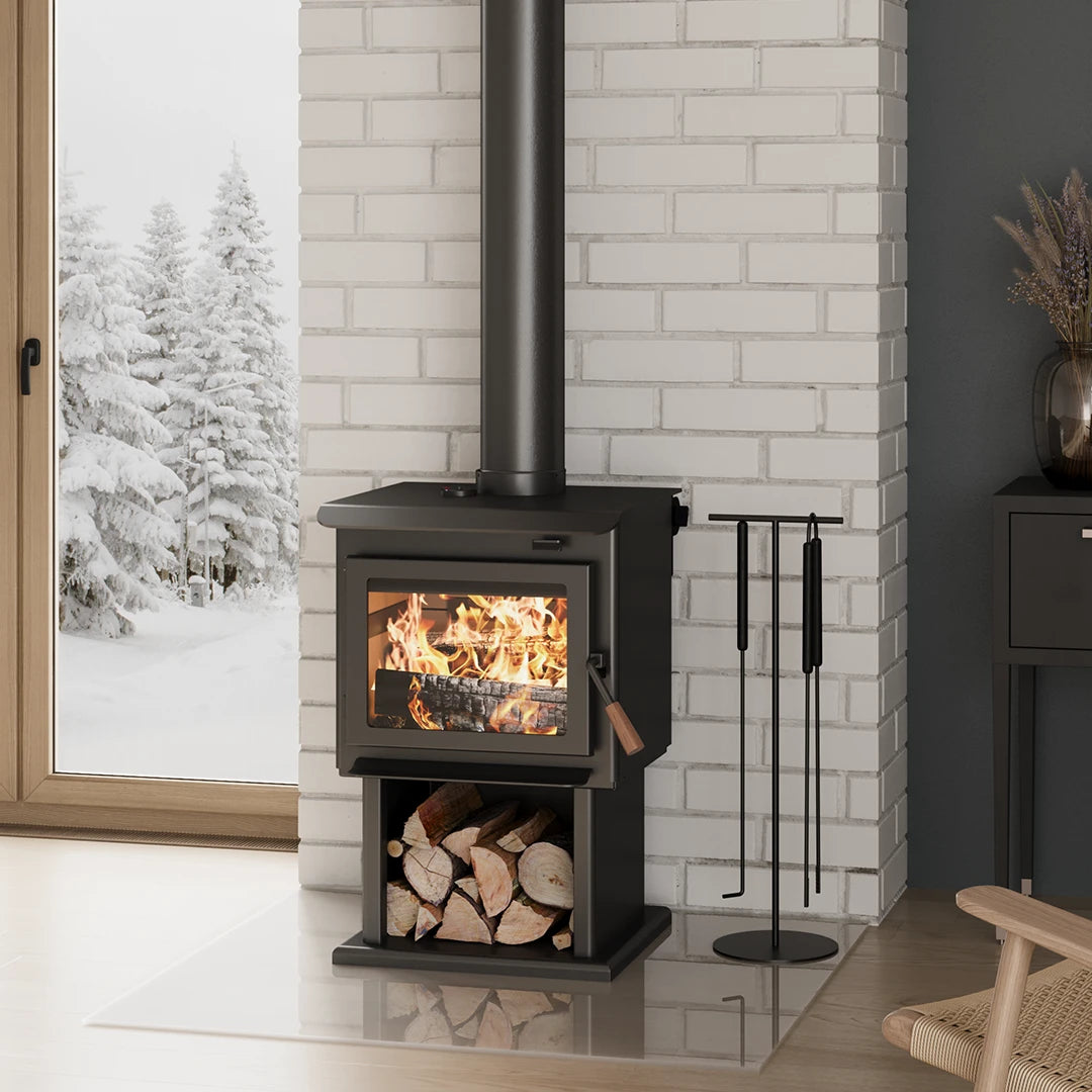 Small Wood-burning stove by Blaze King with fire lit inside, surrounded by a snowy landscape outside a window, wih wood logs stored below.